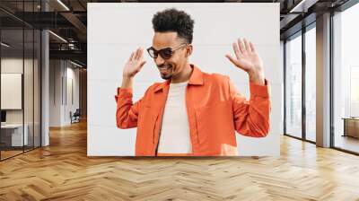 Brunet curly African man in white t-shirt, orange jacket and sunglasses rises hands up and smiles near orange wall outside. Wall mural