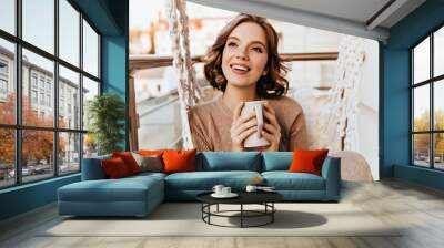 Carefree girl with brown makeup drinking tea at balcony. Photo of pleasant brunette woman in knitted dress enjoying coffee. Wall mural