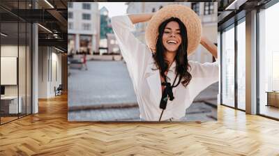 Charming european girl in white blouse expressing happiness in summer day. Outdoor photo of enthusiastic lady in glamorous hat standing on city background. Wall mural