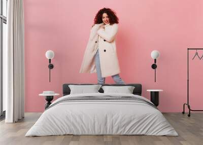 Charming girl with red lipstick posing on pink background. Portrait of curly woman in white wool coat and light jeans Wall mural