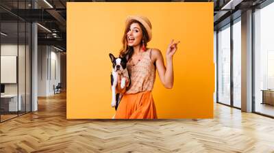 Charming red-haired girl in vintage hat holding french bulldog. Indoor photo of amazed white woman posing on yellow background with puppy. Wall mural