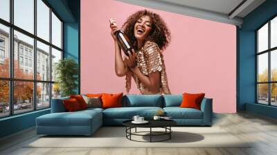 Charming woman with brunette curly hairstyle in shiny stylish dress laughing and holding bottle with wine on isolated backdrop.. Wall mural