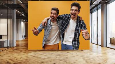 Cheerful brunet happy men in white t-shirts and blue checkered shirts point at camera and laugh on isolated orange background. Wall mural