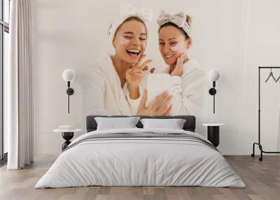 Cheerful caucasian young women after shower carry out skin care procedures applying patches under eyes in spa salon. Ladies wear white robes and headbands. Beauty and youth concept Wall mural