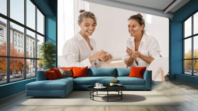 Cheerful caucasian young women apply face lotion laughing sitting in light room. Blondes with gathered hair wear white shirts. Skin care and hydration concept Wall mural