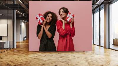 Cheerful curly girl in polka dot black dress smiling, holding gift box and posing with bright lady in red clothes. Wall mural