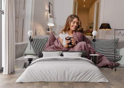 Cheerful girl sitting on couch with blanket and cushions and smiling. Spectacular brunette lady laughing, while drinking coffee under plaid. Wall mural