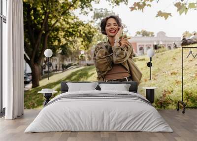 Cheerful woman with short curly hair in white pants with belt smiling outside. Trendy lady in denim olive jacket laughing outdoors.. Wall mural