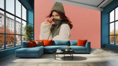 Cheerful young caucasian girl looking into cameras covers her face with collar of sweater on pink background. Brunette with wavy hair wears grey hat, sweater and white jacket.  Wall mural