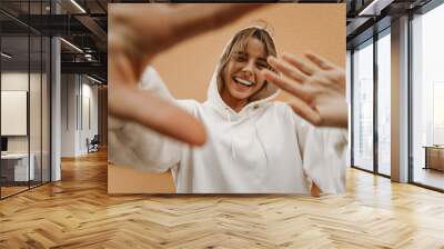 Cheerful young fair-skinned lady hiding from paparazzi hiding behind her hands against background of wall. Blonde with perfect smile wears white sweatshirt. Freedom concept Wall mural