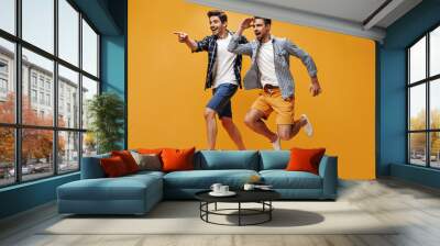 Cheerful young happy men in colorful shorts and checkered blue shirts point into distance, smile and jump on orange background. Wall mural