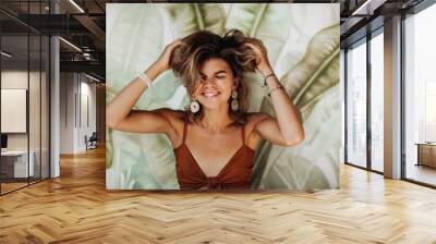 Cheerful young woman with beautiful tan smiles widely and closes eyes. Portrait of emotional happy girl in brown top laughing on background of palm leafs Wall mural