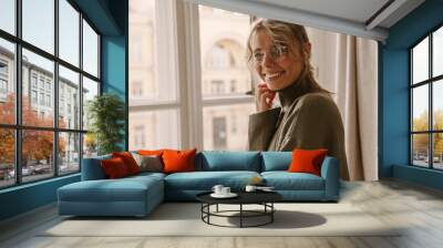 Close-up of cheerful young caucasian lady looking at camera smiling with teeth while standing by window. Attractive blonde in translucent glasses, grey sweater. Mood and good life concept. Wall mural