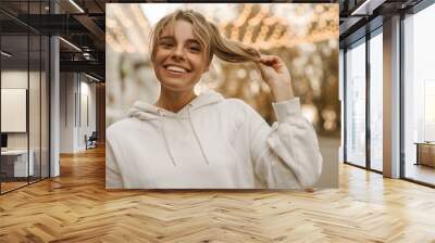 Close-up of young caucasian blonde with gathered hair holding ends of her hair on blurred background. Smiling beauty in white sweatshirt smiles with teeth. Concept enjoying moments Wall mural