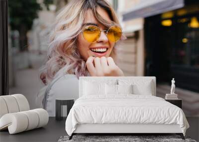 Close-up portrait of attractive joyful girl with amazing smile wears stylish yellow sunglasses and white shirt. Stunning curly young lady with blonde hair excitedly posing on the street. Wall mural