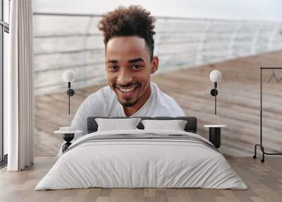 Close-up portrait of happy young dark-skinned man in white sport long-sleeved t-shirt sincerely smiling near sea. Wall mural