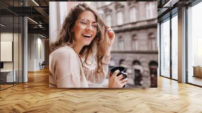 Close-up portrait of laughing brunette girl in beige sweater drinking coffee on city background. Lovable lady standing near window and looking at beautiful building with cup of tea. Wall mural