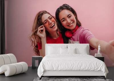 Close-up shot of two interracial young women taking vivid selfie against pink background. Brunette asian with headphones and blonde ukrainian in sunglasses are happy together.  Wall mural