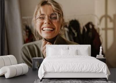 Close-up young caucasian woman laughing hard while screwing up eyes while being indoors. Girl with tousled blonde hair in sweater and glasses smiles broadly. Emotions people, lifestyle concept. Wall mural