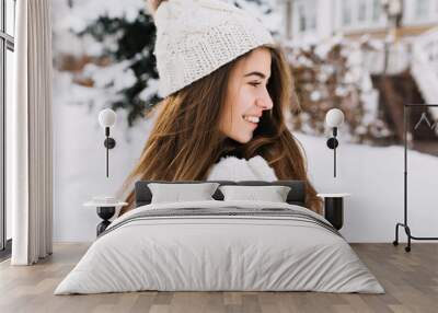 Closeup portrait charming young woman in white woolen gloves, knitted hat, long brunette hair enjoying cold winter weather on street. Smiling to side, true positive emotoins, cheerful mood Wall mural