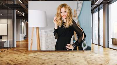 Closeup portrait of young gorgeous woman wearing stylish black dress in cozy studio, touching her wavy hair. Fireplace with firewood, white lamp on background. Wall mural