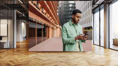 Concerned young african man talking on phone walking down street. Brunette guy with stubble wears shirt in spring. Concept of use Wall mural