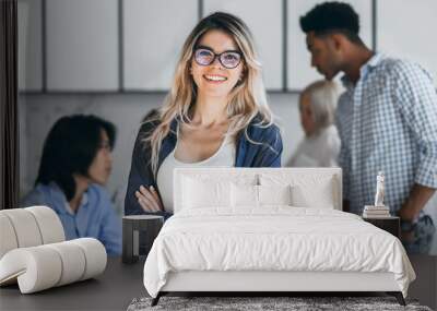 Confident blonde female manager posing with smile after conference with other employees. Asian programmer talking with african freelancer while fair-haired secretary laughing on foreground. Wall mural