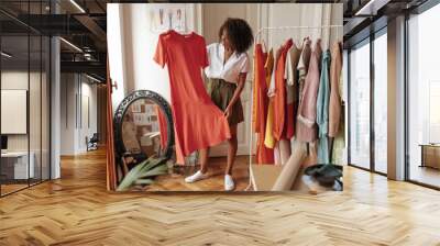 Curly stylish dark-skinned woman in khaki shorts and white blouse holds hanger with long red dress and poses in dressing room. Wall mural