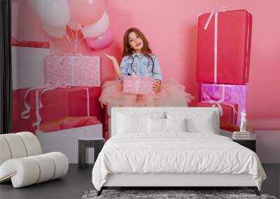 Cute charming little girl in tulle skirt sitting with present on knees suround big giftboxes, balloons on pink background. Happy celebration birthday party in childhood, expressing true emotions Wall mural