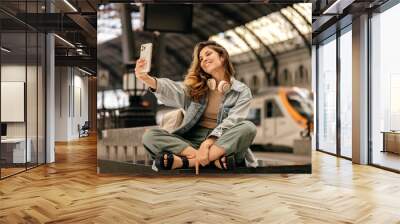 Cute young caucasian blonde woman makes selfie on smartphone sitting at railway station. Girl wears casual clothes. Technology concept, life Wall mural