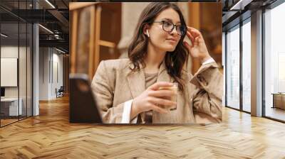 Cute young caucasian girl spends time in street cafe using digital tablet. Brunette wears glasses for vision and beige jacket. Weekend enjoyment concept Wall mural