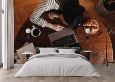 Dark-haired girl fell asleep on wooden table while working in laptop. There is cup of coffee and croissants on table Wall mural