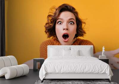 Dark-haired young woman with big suprised eyes posing on orange background. Studio shot of elegant caucasian girl in knitted clothes expressing amazement during photoshoot. Wall mural