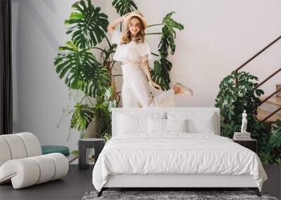 Ecstatic blonde girl in white pants funny dancing at home posing in front of big plant. Indoor portrait of jumping young woman in hat and glasses happy smiling next to stairs. Wall mural