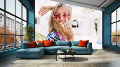 Ecstatic girl wears pink sunglasses expressing happiness in summer day. Outdoor photo of wonderful tanned woman posing with pretty smile in sunny morning. Wall mural