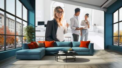 Elegant business-lady in trendy black jacket holding laptop and smiling. Portrait of cheerful blonde secretary and tall african office worker on background. Wall mural