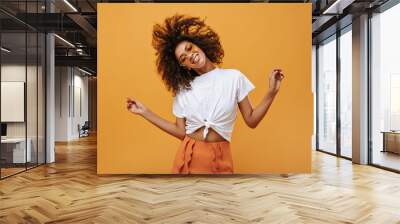 Emotional curly girl in light t-short playing her hair on isolated backdrop. Joyful woman in glasses poses on yellow background.. Wall mural