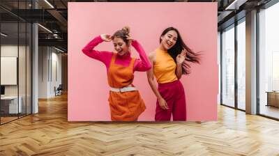 Energetic young two interracial girls dancing side by side and waving hands in studio. Models with light brown and black hair in light spring orange and pink clothes move to music with smile on face. Wall mural