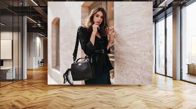 Engaging young lady with brunette wavy hair, bright makeup, dark modern outfit and black bag on shoulder, thoughtfully posing near beige sunlit wall outdoors Wall mural