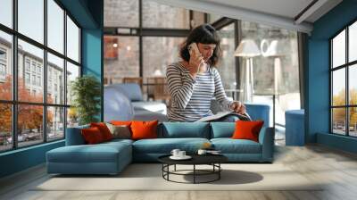 European business woman talking on cellphone and look at workbooks. Sitting in coworking place, wearing blue stripes shirt, jeans. Freelance work concept  Wall mural