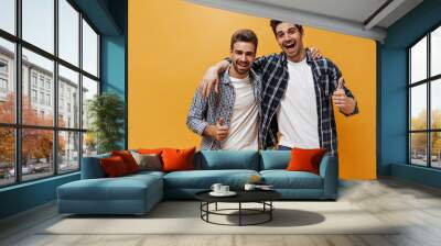 Excited brunet men in checkered shirts, colorful pants and white t-shirts smile, show thumbs up and pose on orange background. Wall mural