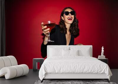 Excited curly woman in sunglasses and festive black dress holds cocktail glass and laughs on red background. Wall mural