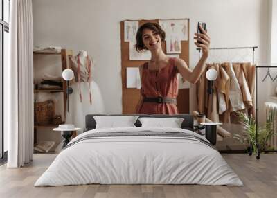 Excited short-haired young woman in trendy linen dress with black belt smiles, poses in office of fashion designer and holds phone. Wall mural