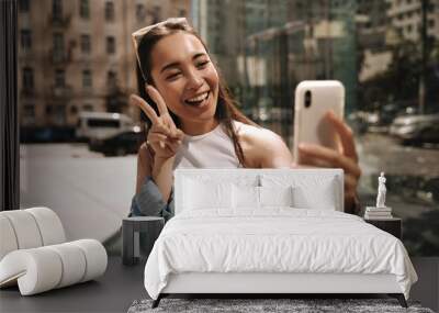 Excited young asian girl in white t-shirt using smartphone makes selfie showing two fingers on street. Cute lady takes pictures of herself and looks at phone screen. Wall mural