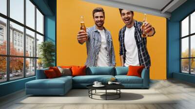 Excited young men in white trendy t-shirts and checkered shirts rejoice and hold beer bottles on orange isolated background. Wall mural