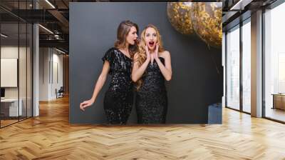 Fascinating blonde girl in luxury attire spending time at party with best friend. Attractive fair-haired girl in black dress posing with surprised face on dark background during event. Wall mural