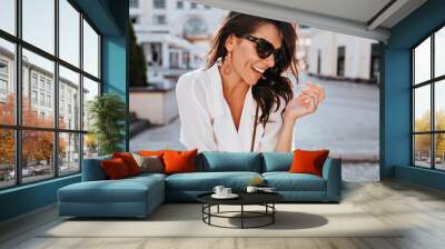 Fascinating female model in earrings laughing in good day. Attractive brunette woman in sunglasses having fun outdoor. Wall mural