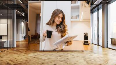 Fascinating female model with light-brown hair reading journal in her kitchen. Indoor photo of pretty smiling girl with cup of tea holding magazine. Wall mural