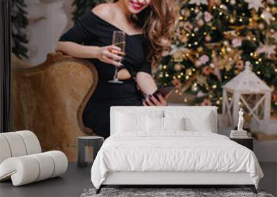 full-length photo of girl in elegant total black outfit, gazing at messages in phone. Brunette sits on beautiful soft sofa on background of Christmas tree Wall mural