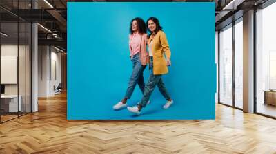 Full-length studio shot of two trendy girls walking on blue background. Smiling african and latin women posing in jeans. Wall mural